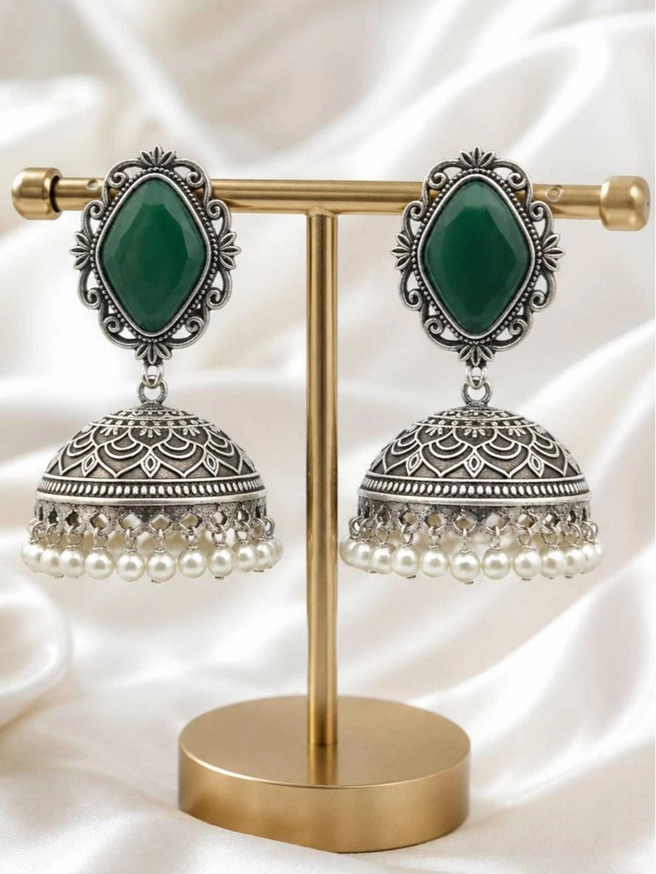 Pair of oxidised silver jhumka earrings with green stone tops and white pearl drops displayed on a gold earring stand