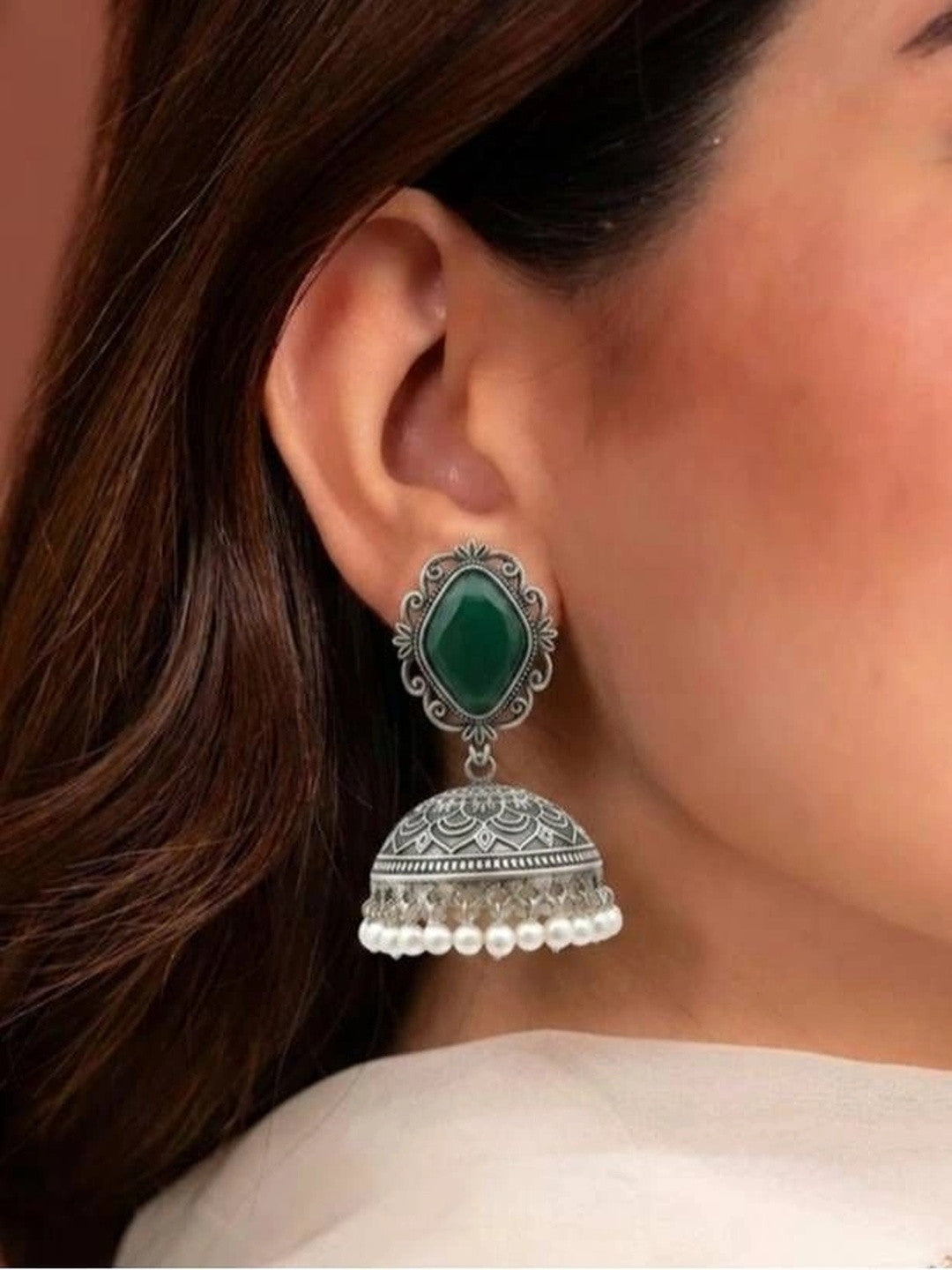 Close-up side profile of oxidised silver green stone jhumka earring worn on a womans ear against pink background
