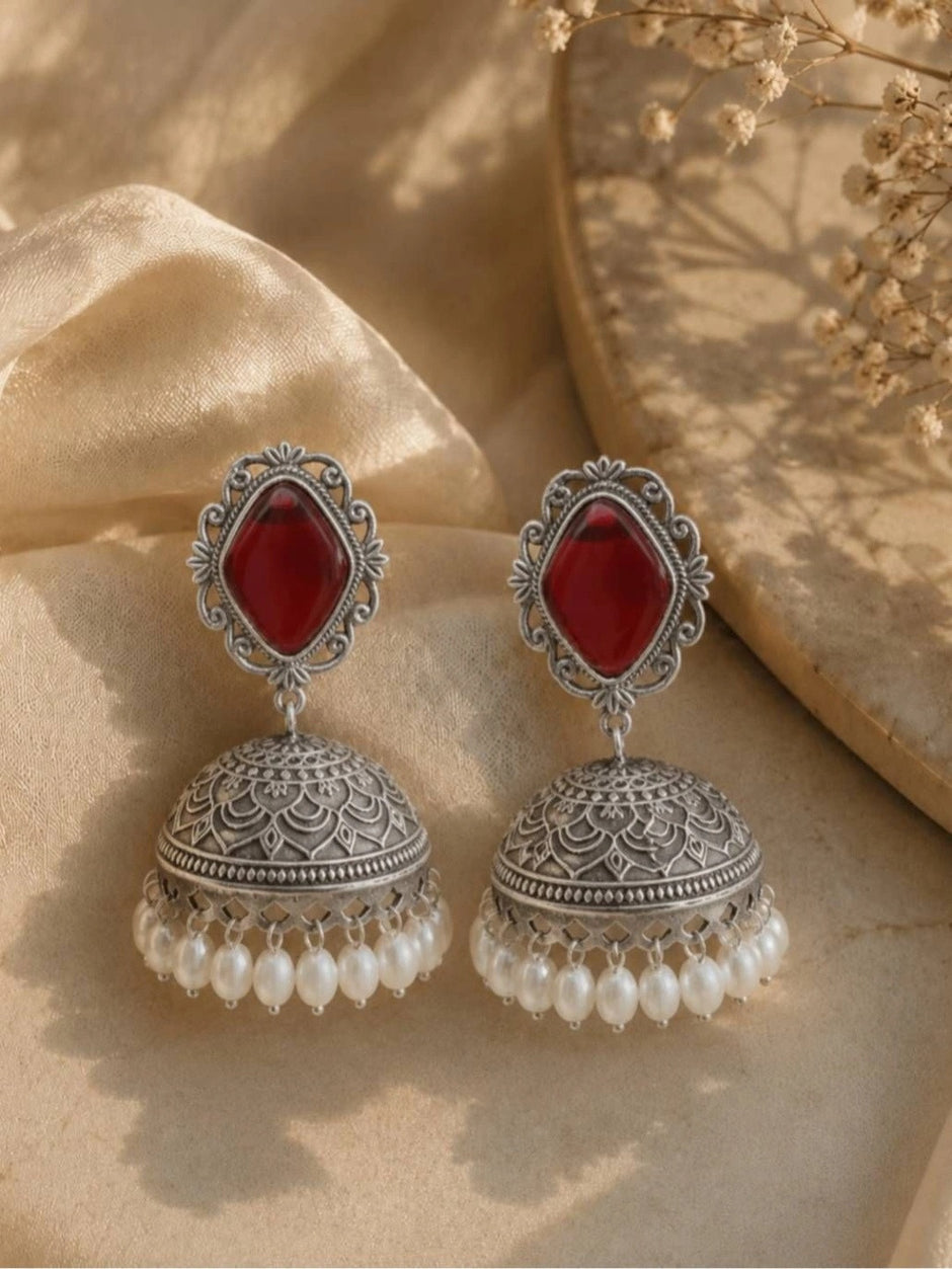 Pair of oxidised silver jhumka earrings with red stone studs and white pearl drop fringe on a cream fabric background