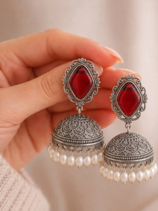 Close-up of oxidised silver jhumka earrings with red glass stone held in a woman's hand showing intricate dome engraving and pearl drops