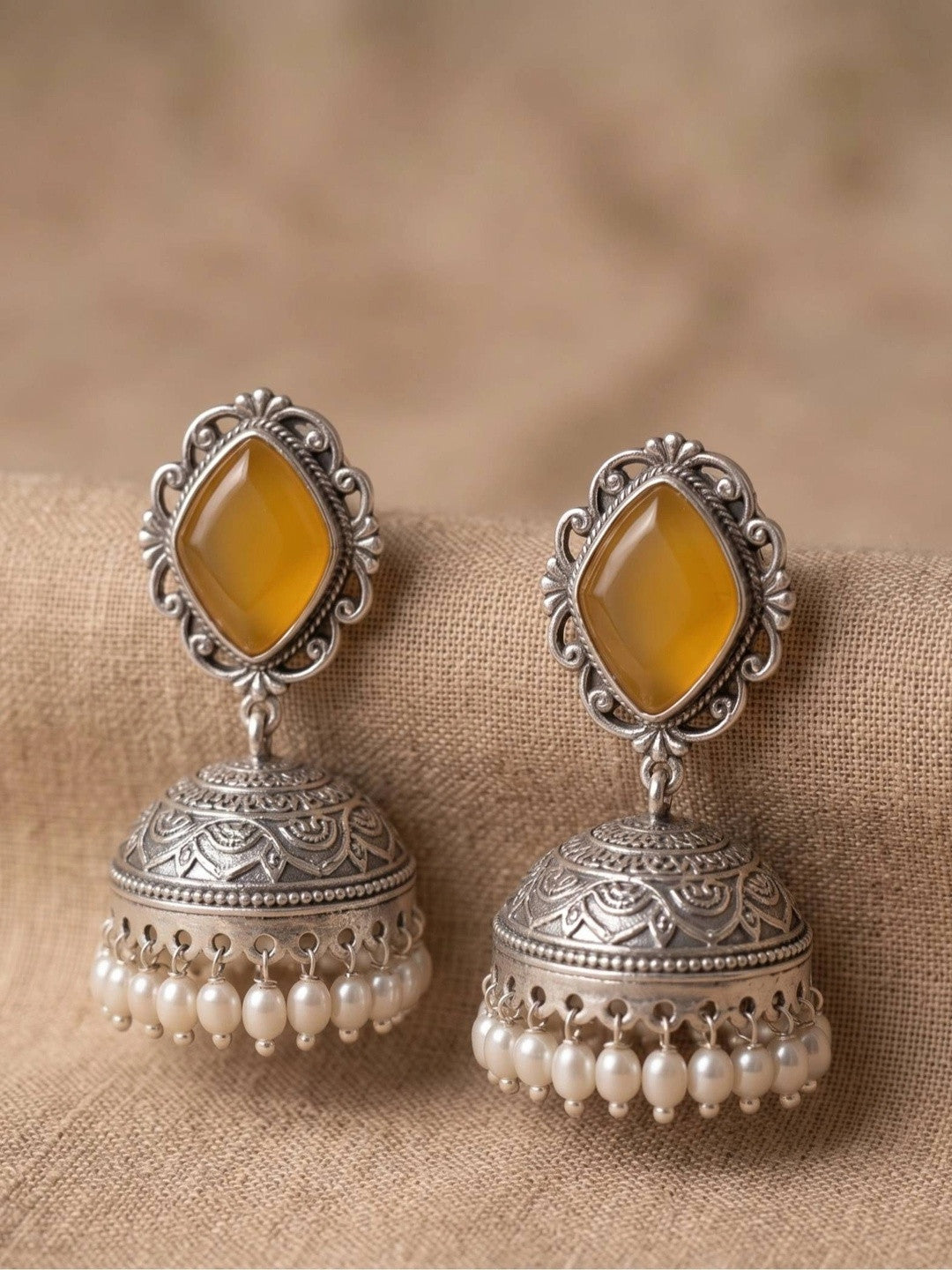Pair of oxidised silver jhumka earrings with yellow diamond-shaped stones and pearl drop fringe, placed on beige fabric