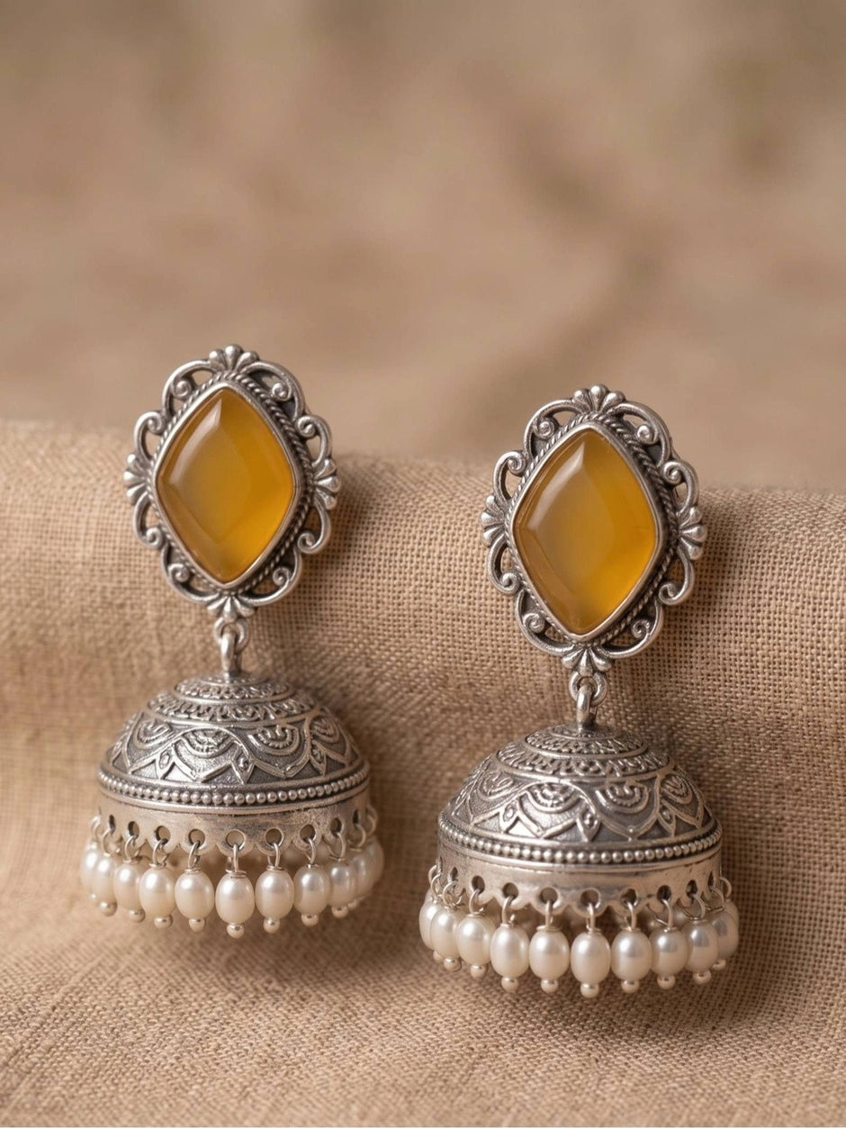 Pair of oxidised silver jhumka earrings with yellow diamond-shaped stones and pearl drop fringe, placed on beige fabric