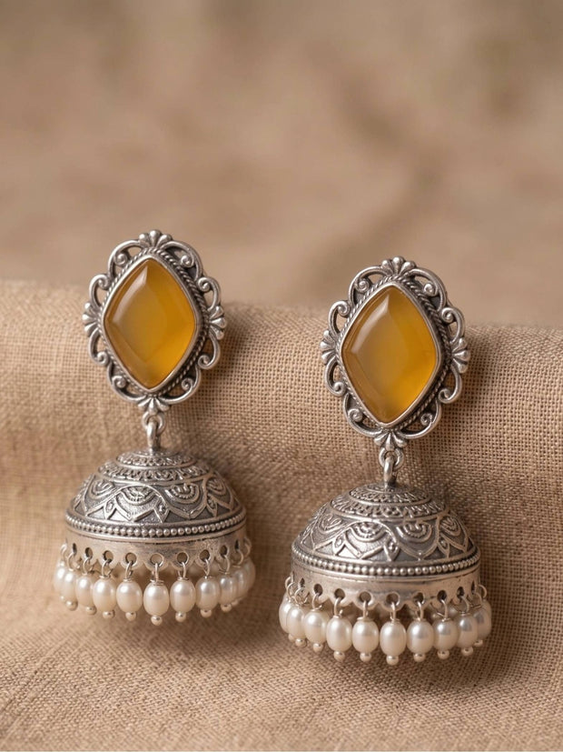 Pair of oxidised silver jhumka earrings with yellow diamond-shaped stones and pearl drop fringe, placed on beige fabric