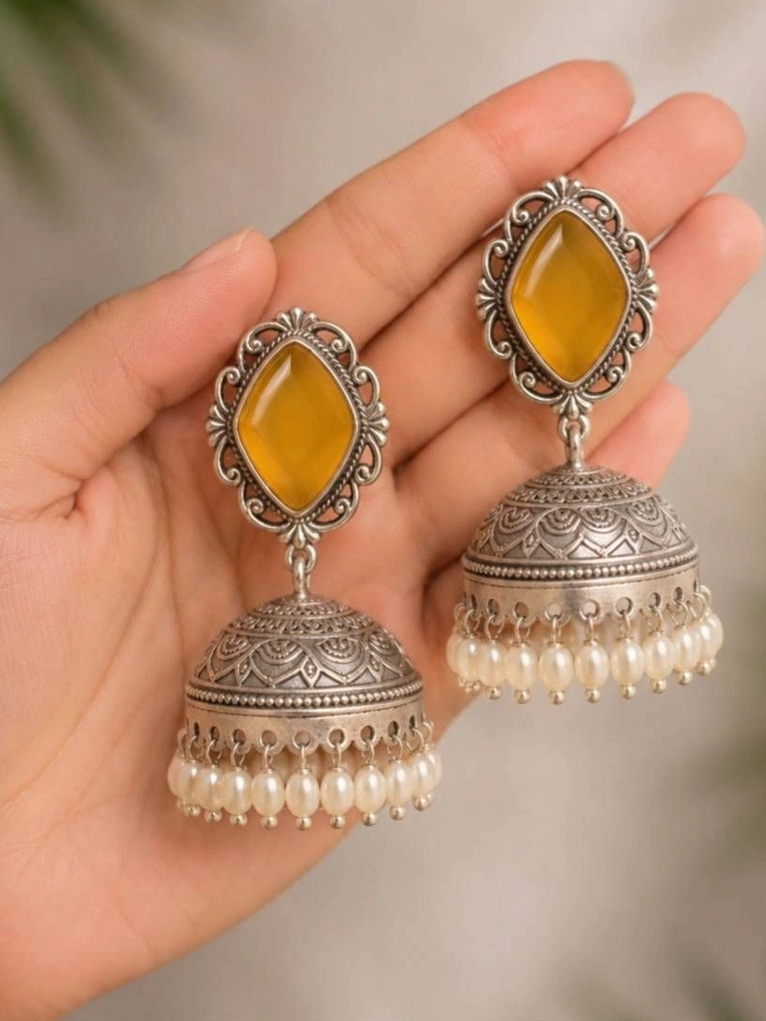Close-up of two oxidised silver jhumka earrings with yellow stones and pearl drops resting on an open palm