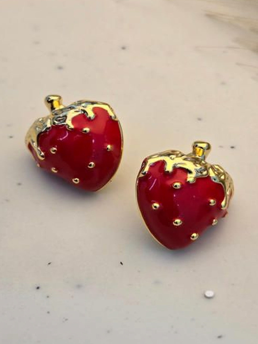 Pair of red enamel strawberry stud earrings with gold-plated leaves on a white marble surface