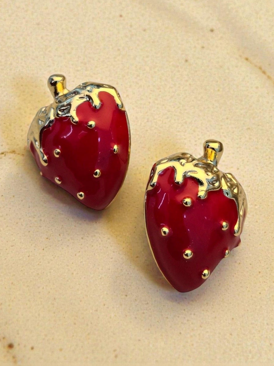 Red enamel strawberry studs with gold-tone detailing resting on a warm sandy beige background