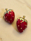 Red enamel strawberry studs with gold-tone detailing resting on a warm sandy beige background