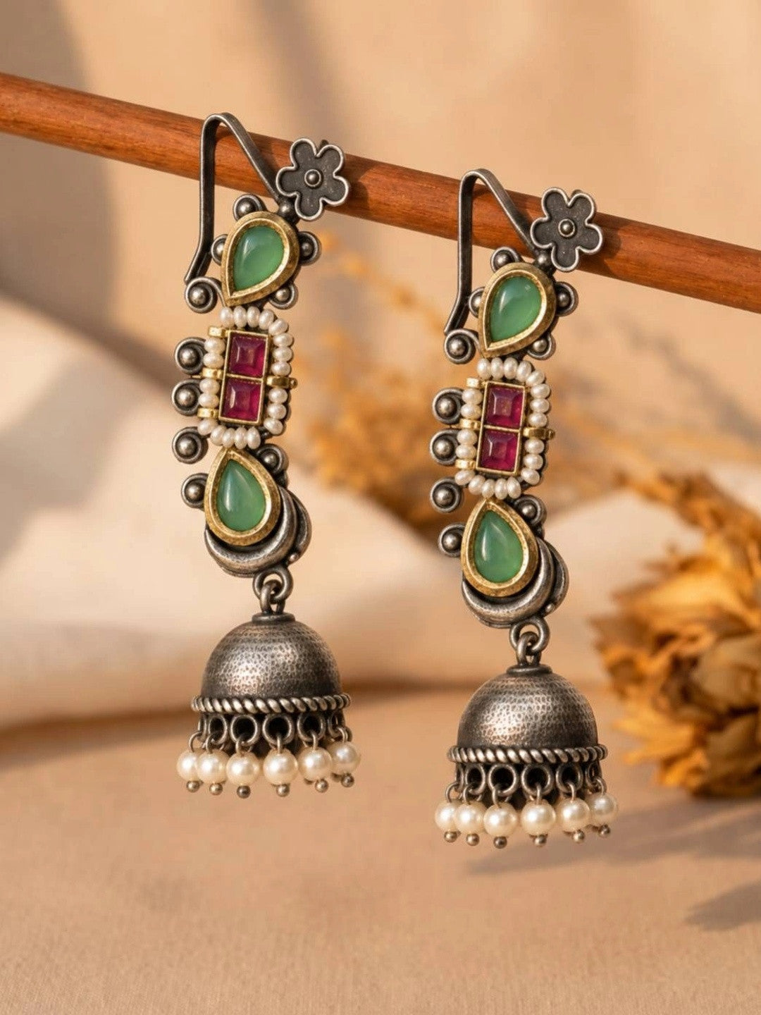 Pair of oxidised silver ear cuff jhumkas with green teardrop stones and pearl-trimmed red stone centre, hanging on a wooden rod