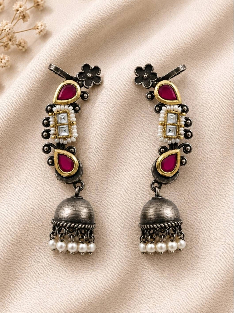 Pair of oxidised silver Kundan ear cuff jhumkas with ruby teardrops and pearl drops laid flat on ivory fabric