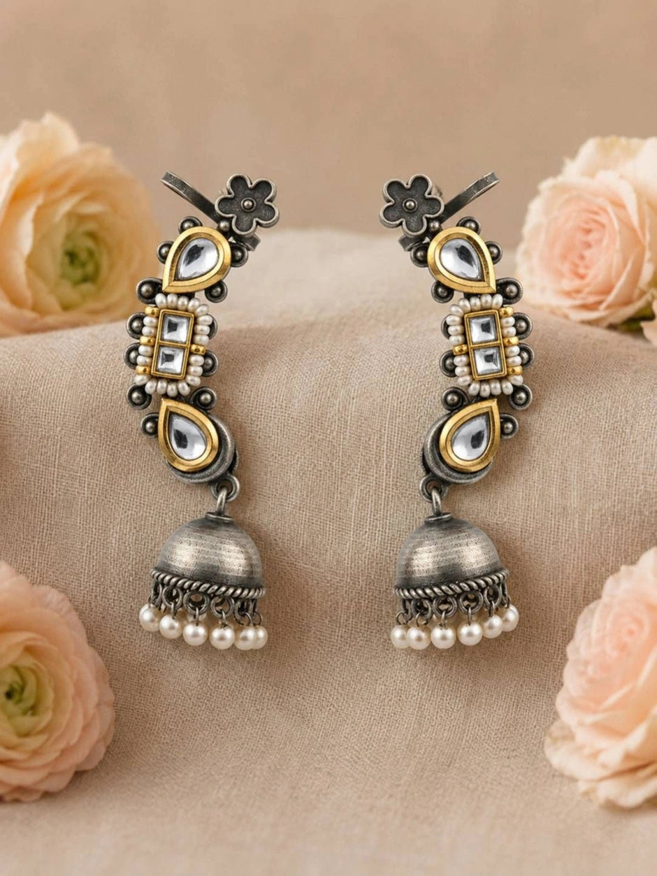 Pair of oxidised dual-tone kundan ear cuff jhumka earrings laid flat on beige fabric surrounded by peach roses