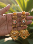 A pair of gold-plated meenakari chandbali earrings held in a hand against green foliage