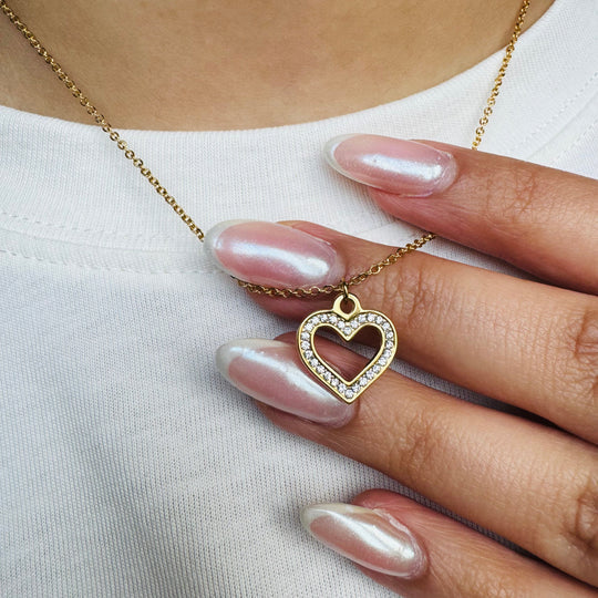Close-up of a hand holding a gold necklace with an open heart-shaped pendant encrusted with white crystals, styled with glossy pearl nails on a white top.