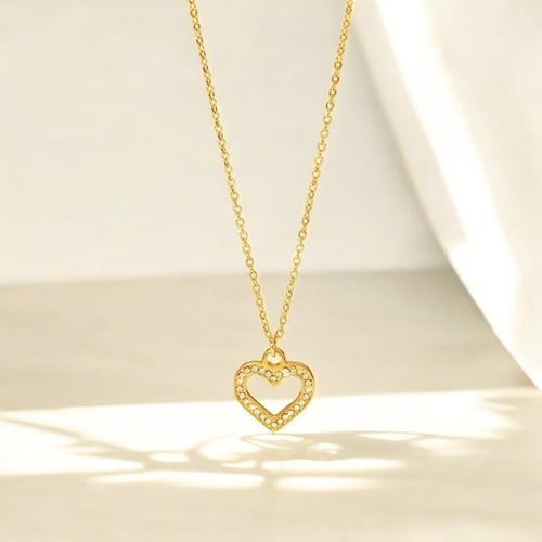 A gold heart-shaped pendant necklace with a hollow center, placed against a soft, draped background.