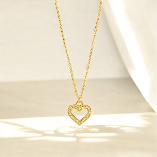 A gold heart-shaped pendant necklace with a hollow center, placed against a soft, draped background.
