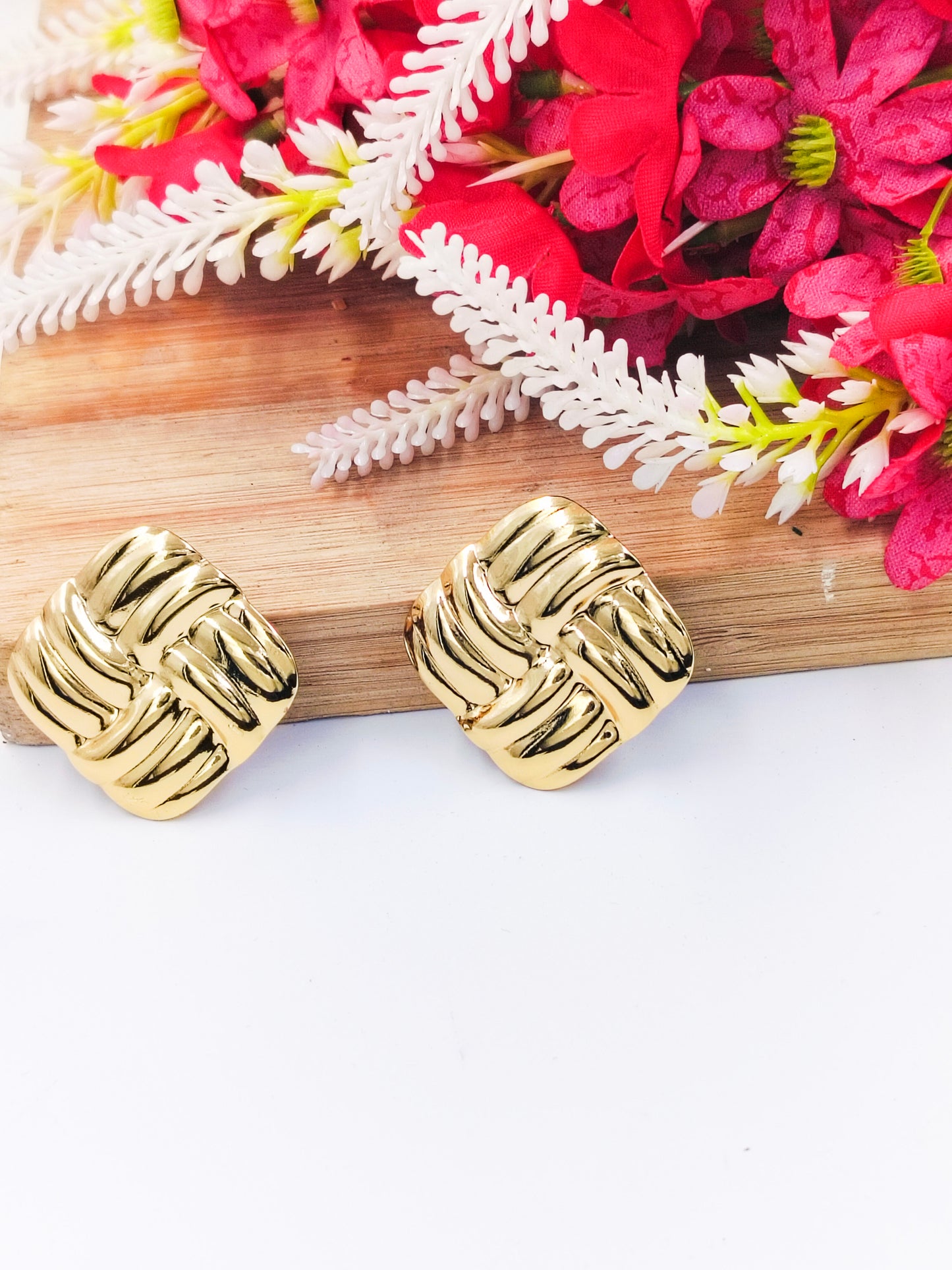 Chic gold square stud earrings with woven texture design, styled with red and white flowers in the background.