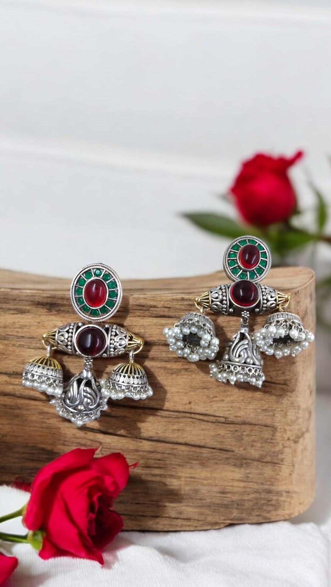 A pair of antique Matrika Mandala Jhumkas earrings with red and green color fusion, displayed on a wooden plank next to a red rose.