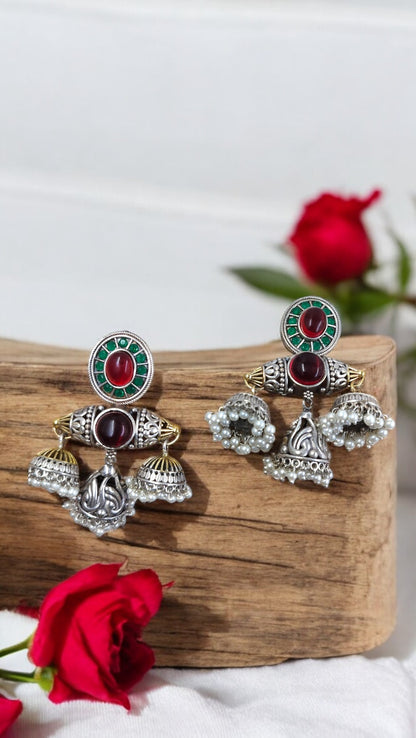 A pair of antique Matrika Mandala Jhumkas earrings with red and green color fusion, displayed on a wooden plank next to a red rose.