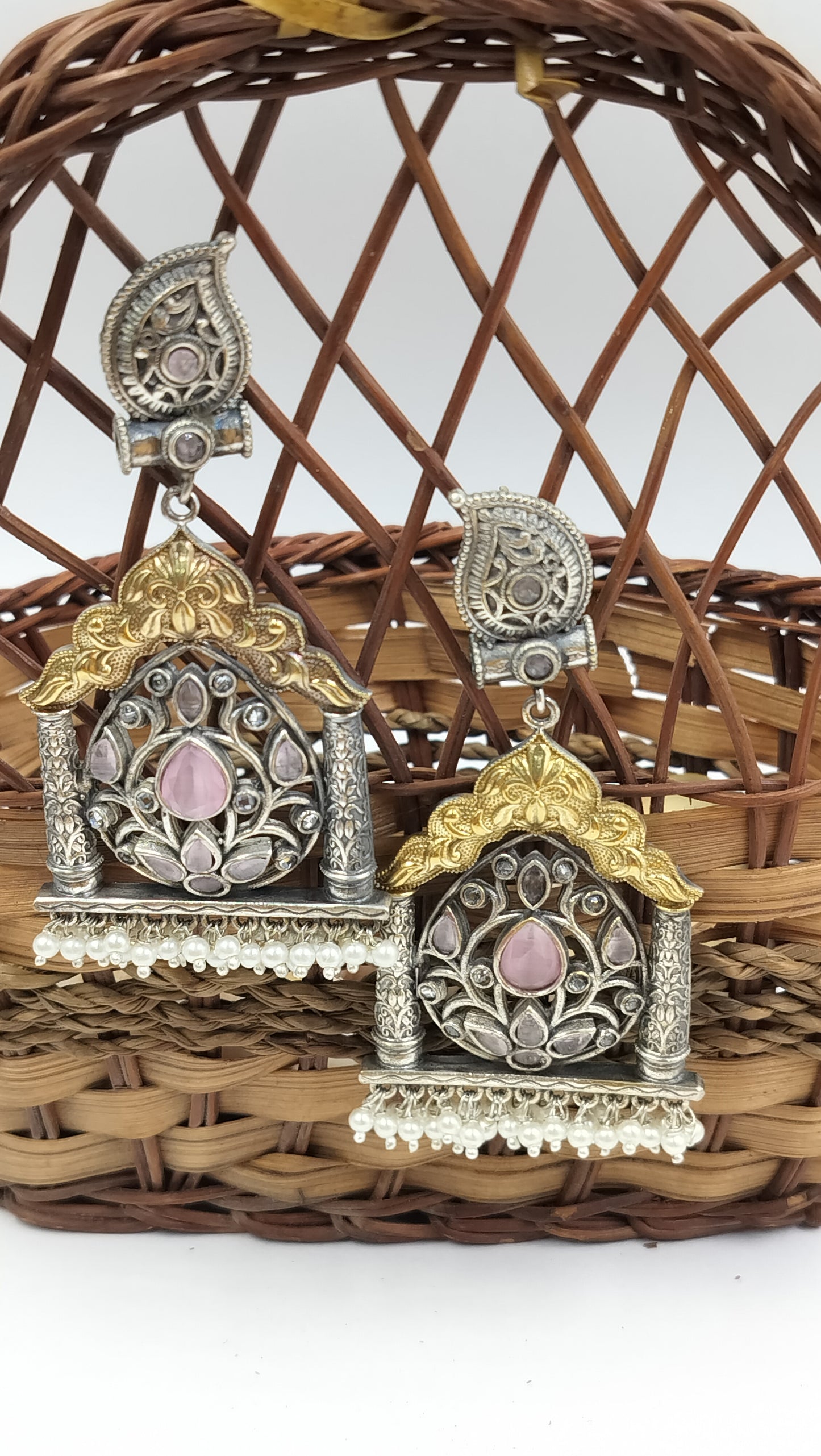 Traditional silver temple earrings with pink gemstone center, floral motifs, and golden arch design, set on wicker basket.