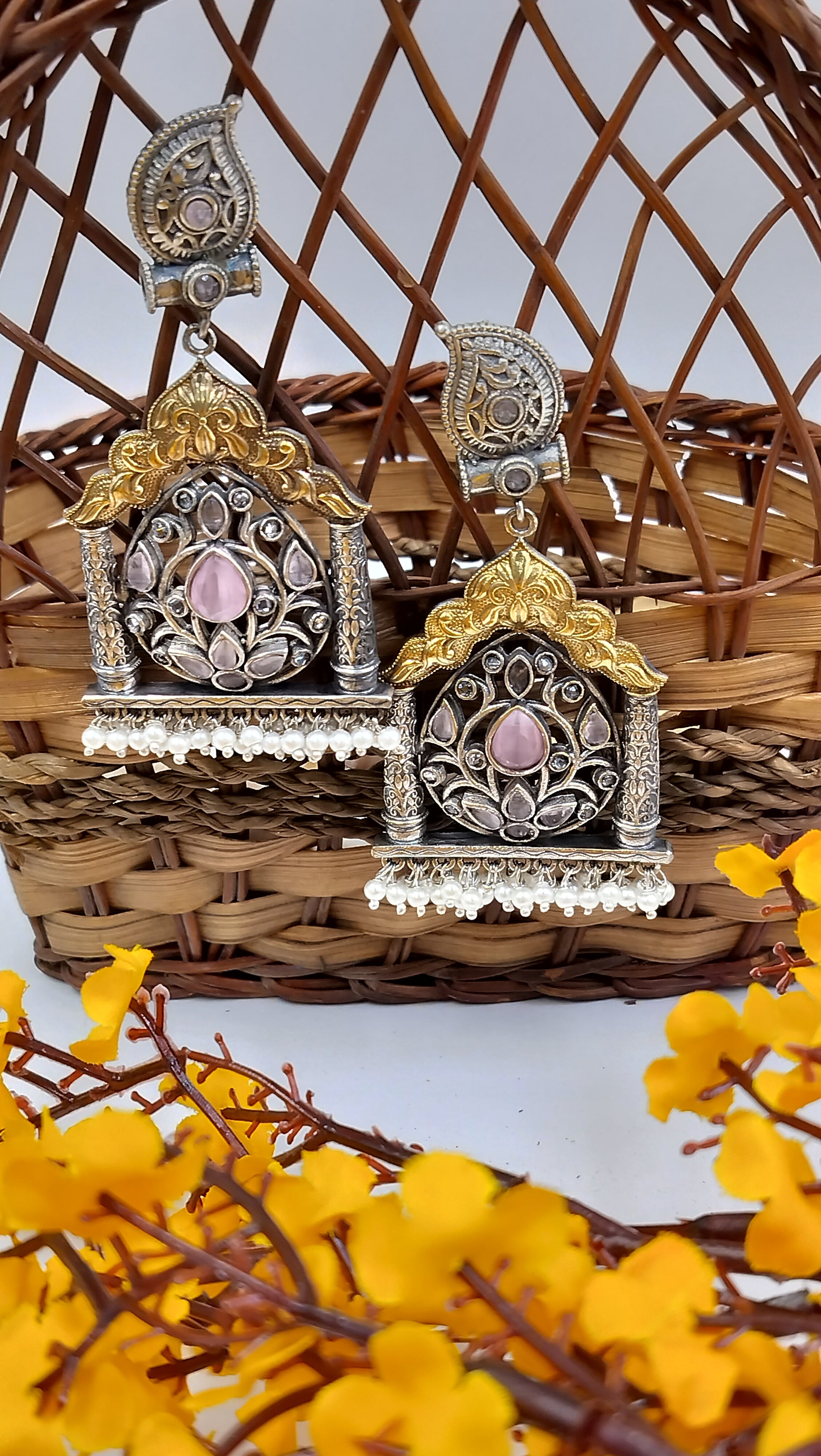 Silver temple-style earrings with pink stone center, floral detailing, and golden top, placed on basket with yellow flowers.