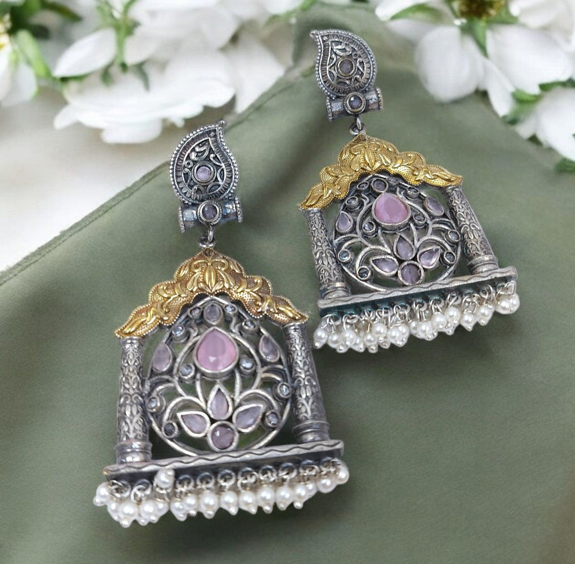 Ornate silver and gold earrings with pink stones on a green fabric background