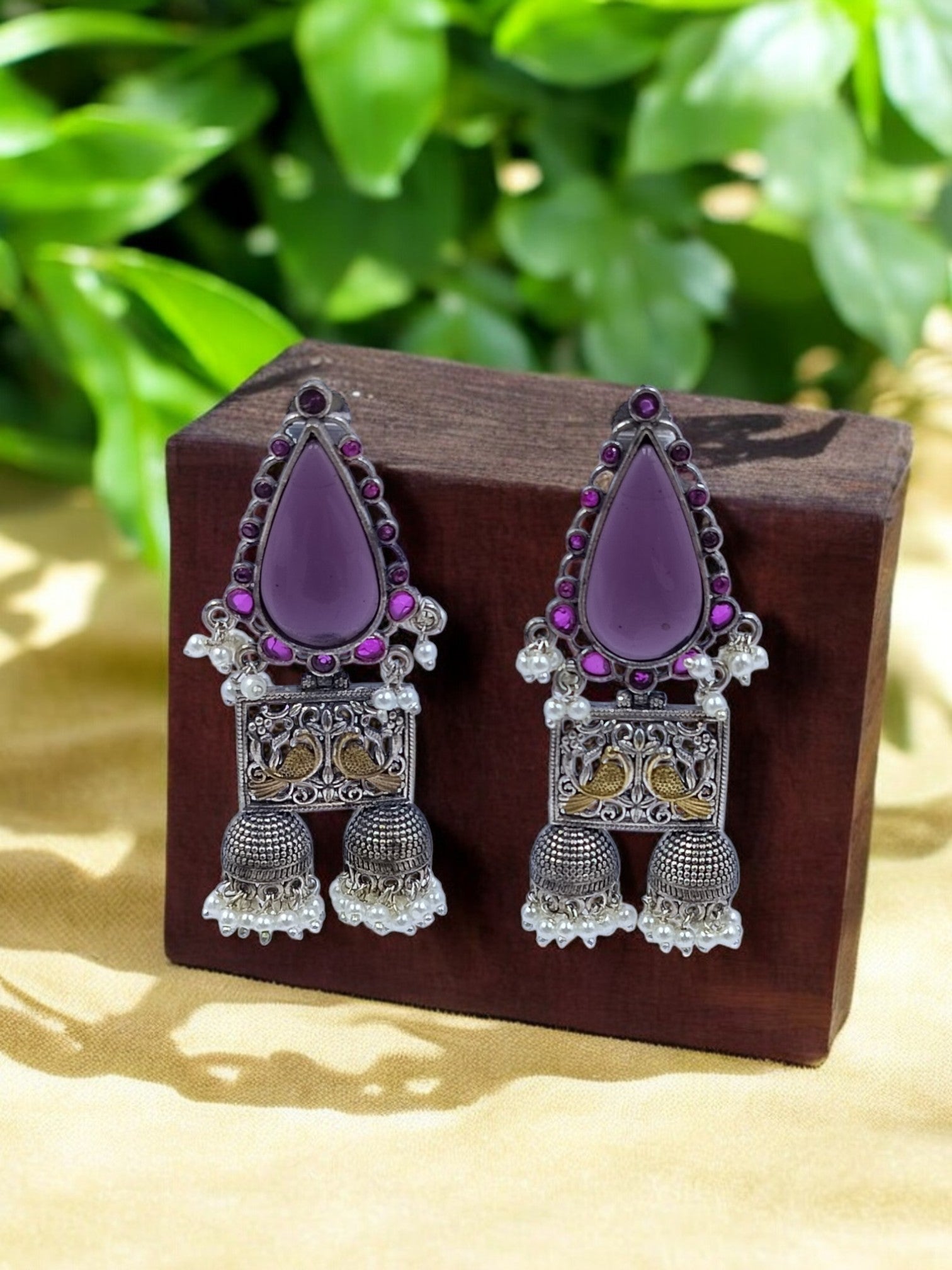 "A pair of deep purple fusion earrings placed on a wooden box, styled with green leaves in the background.