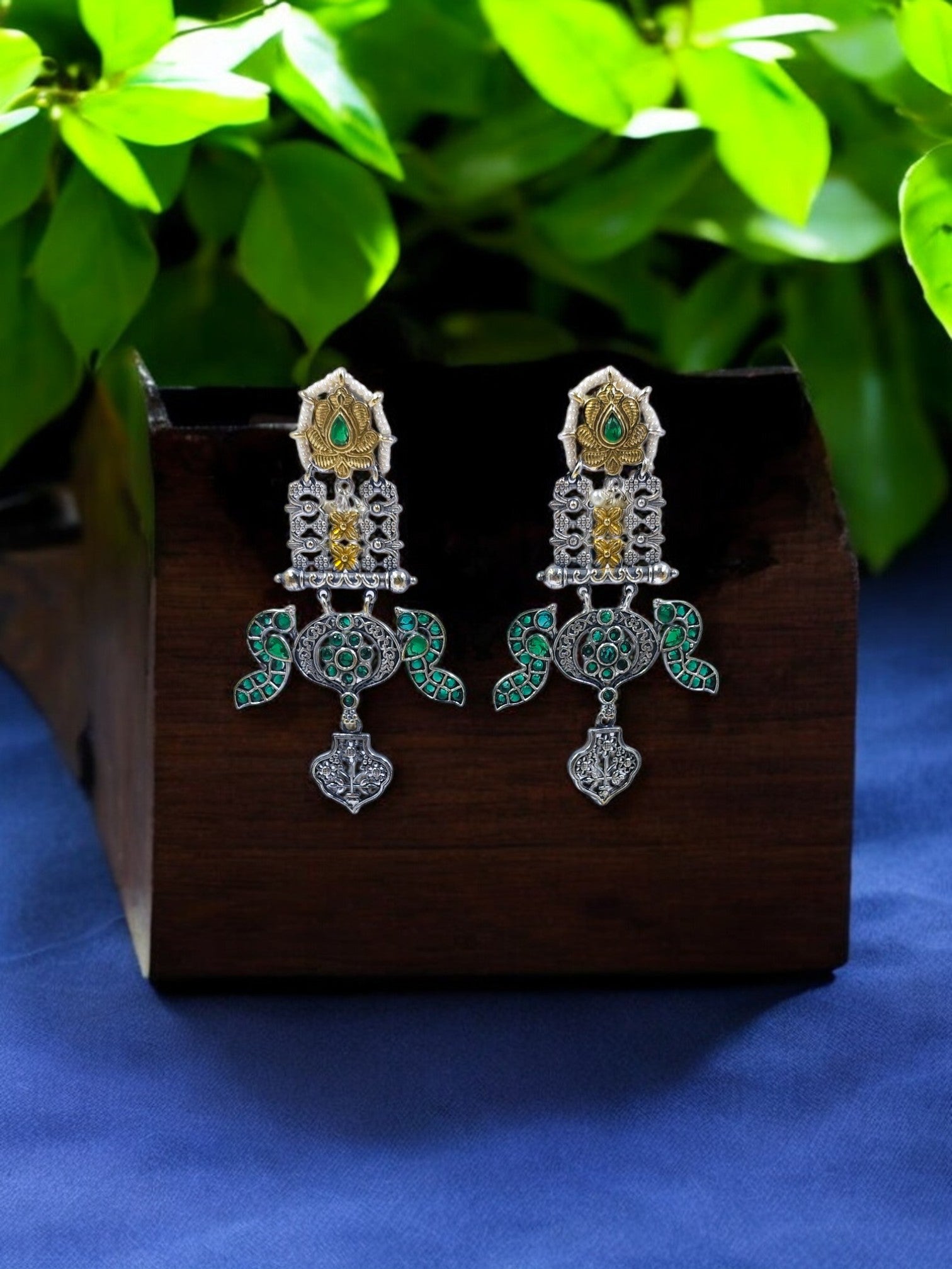 Antique dual-tone earrings with green accents and intricate motifs, styled on a wooden box with leafy backdrop.