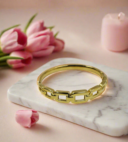 A gold-colored bracelet laid on a white marble surface with pink tulips and a pink candle in the background.