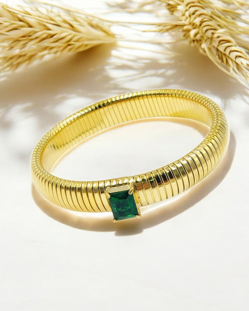 A gold-colored stretchable snake coil elastic bangle bracelet with a square green emerald centerpiece, displayed against a neutral background with a shadow cast over it.