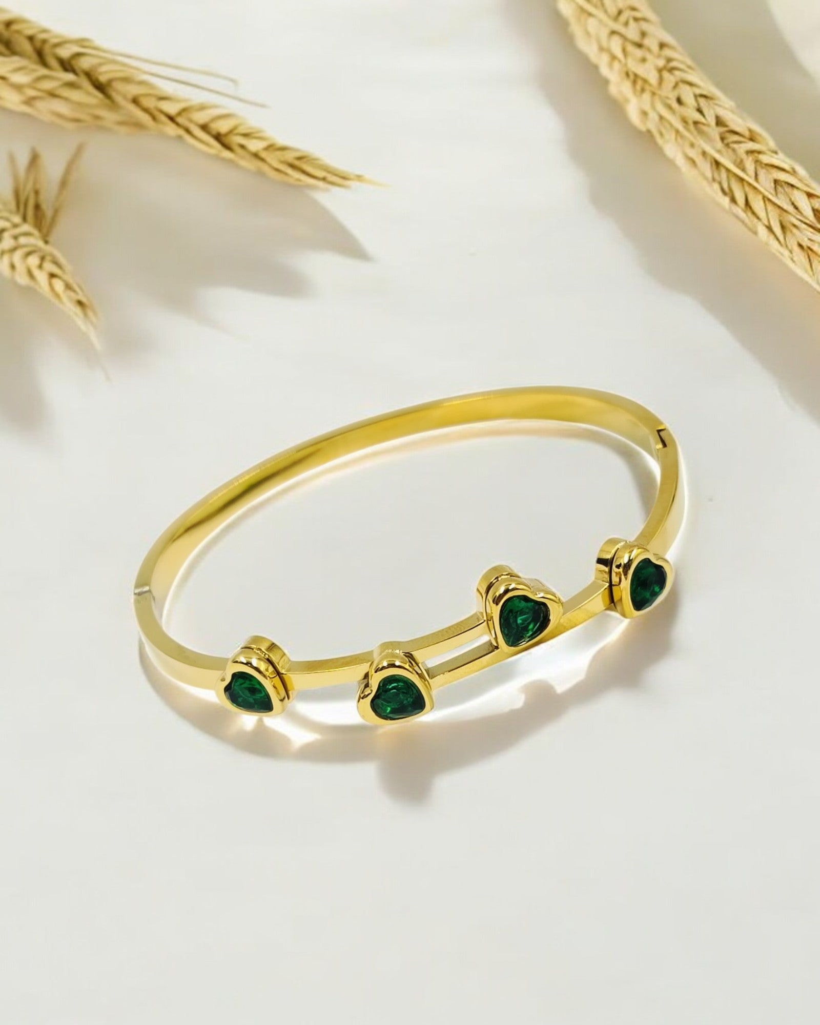 A gold-colored band ring with multiple green heart-shaped stones set on it, displayed against a neutral background with wheat shears.