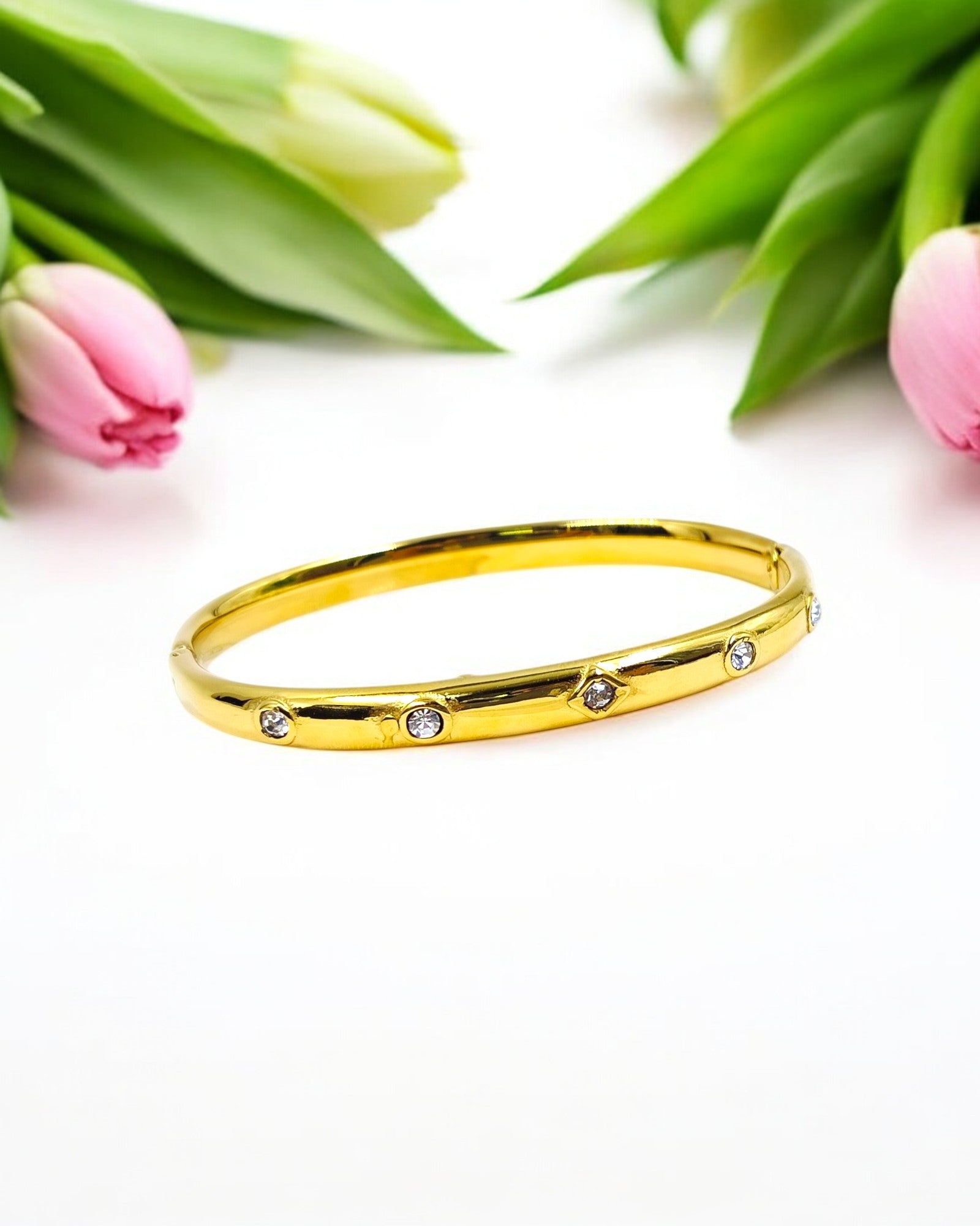 A gold-colored bracelet with diamond-like studs, displayed alongside pink tulips.