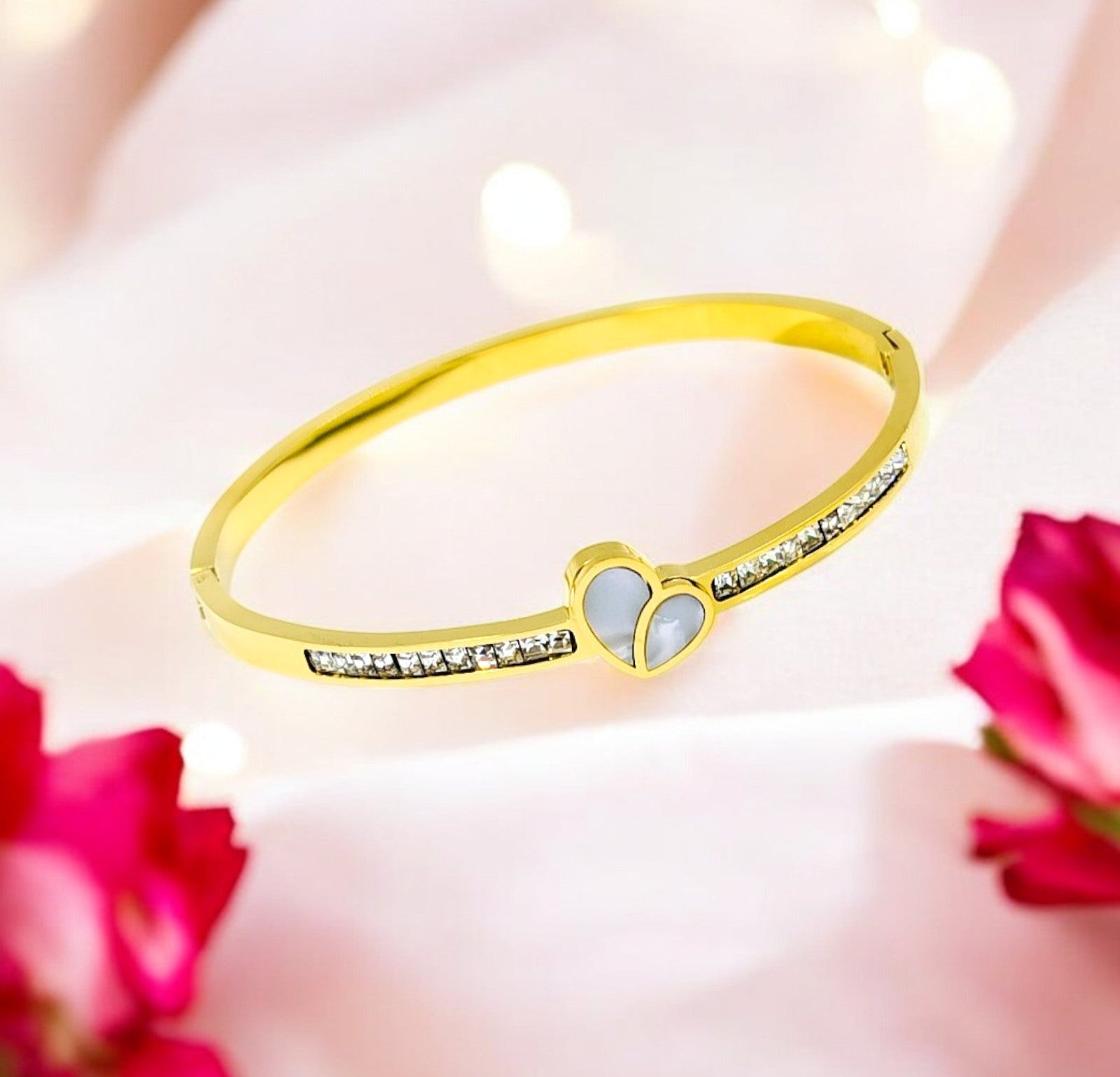 A gold-colored bangle bracelet with a heart-shaped centerpiece and embedded stones, displayed against a floral background.