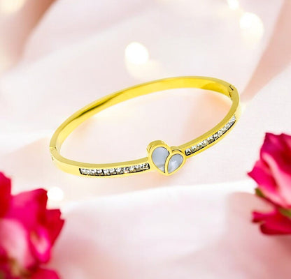 A gold-colored bangle bracelet with a heart-shaped centerpiece and embedded stones, displayed against a floral background.