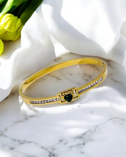 A gold bangle bracelet with a heart-shaped centerpiece and embedded stones, displayed on a white marble surface with yellow flowers in the background.