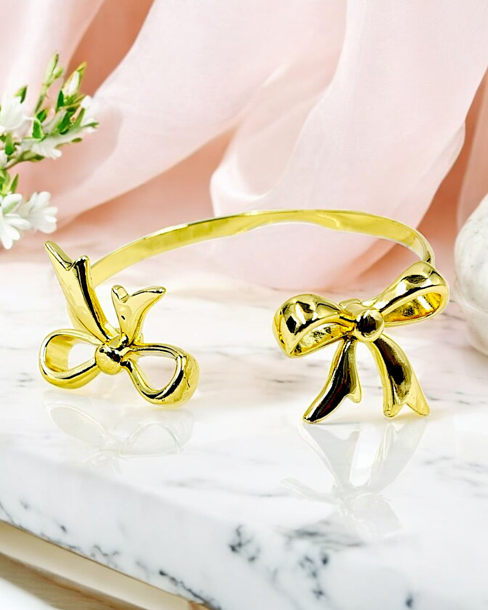 A gold bangle bracelet with bow-shaped accents, displayed on a marble surface with a pink flower in the background.