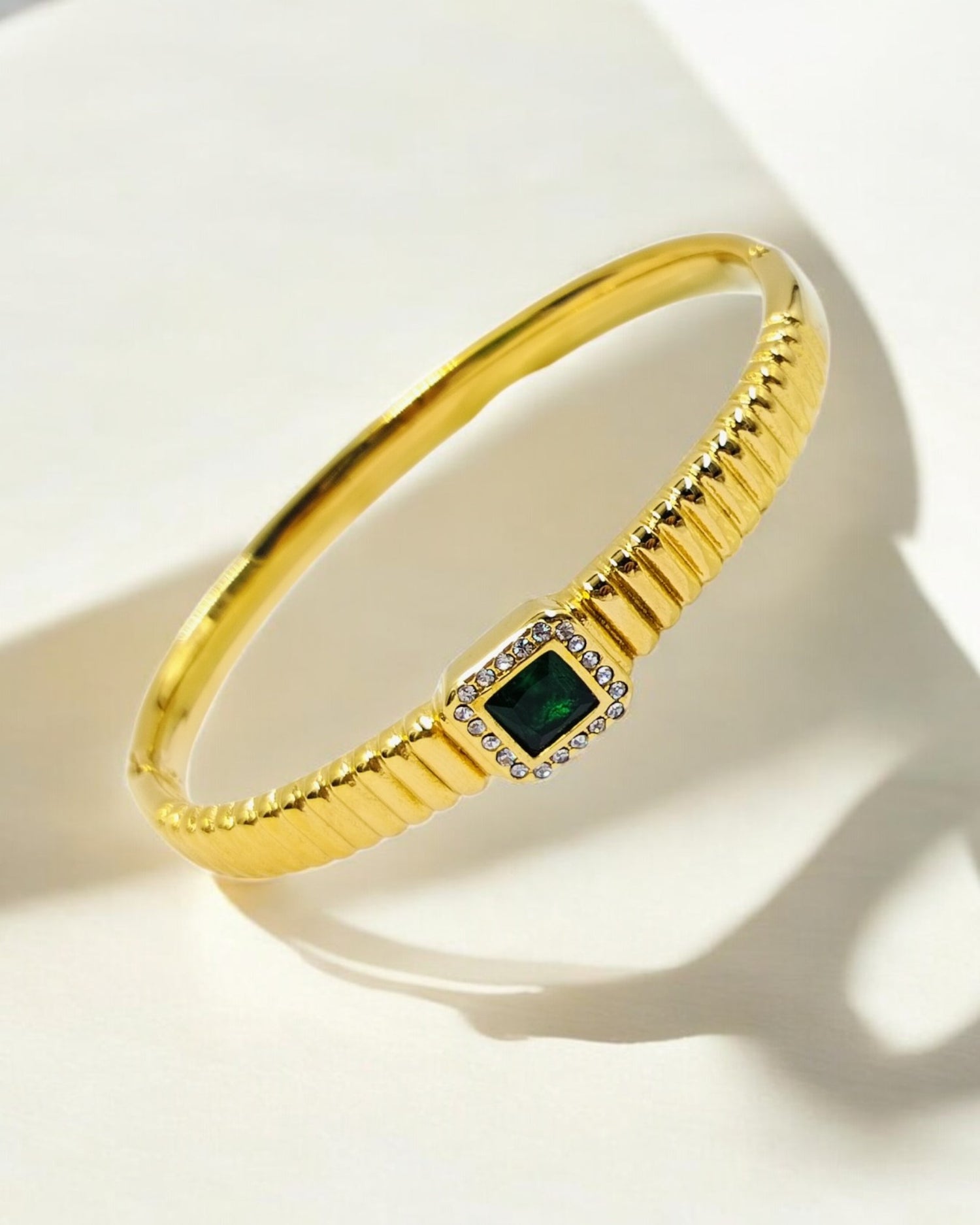 A gold-colored bangle bracelet with a textured band, featuring an emerald centerpiece and diamond-studded frame.