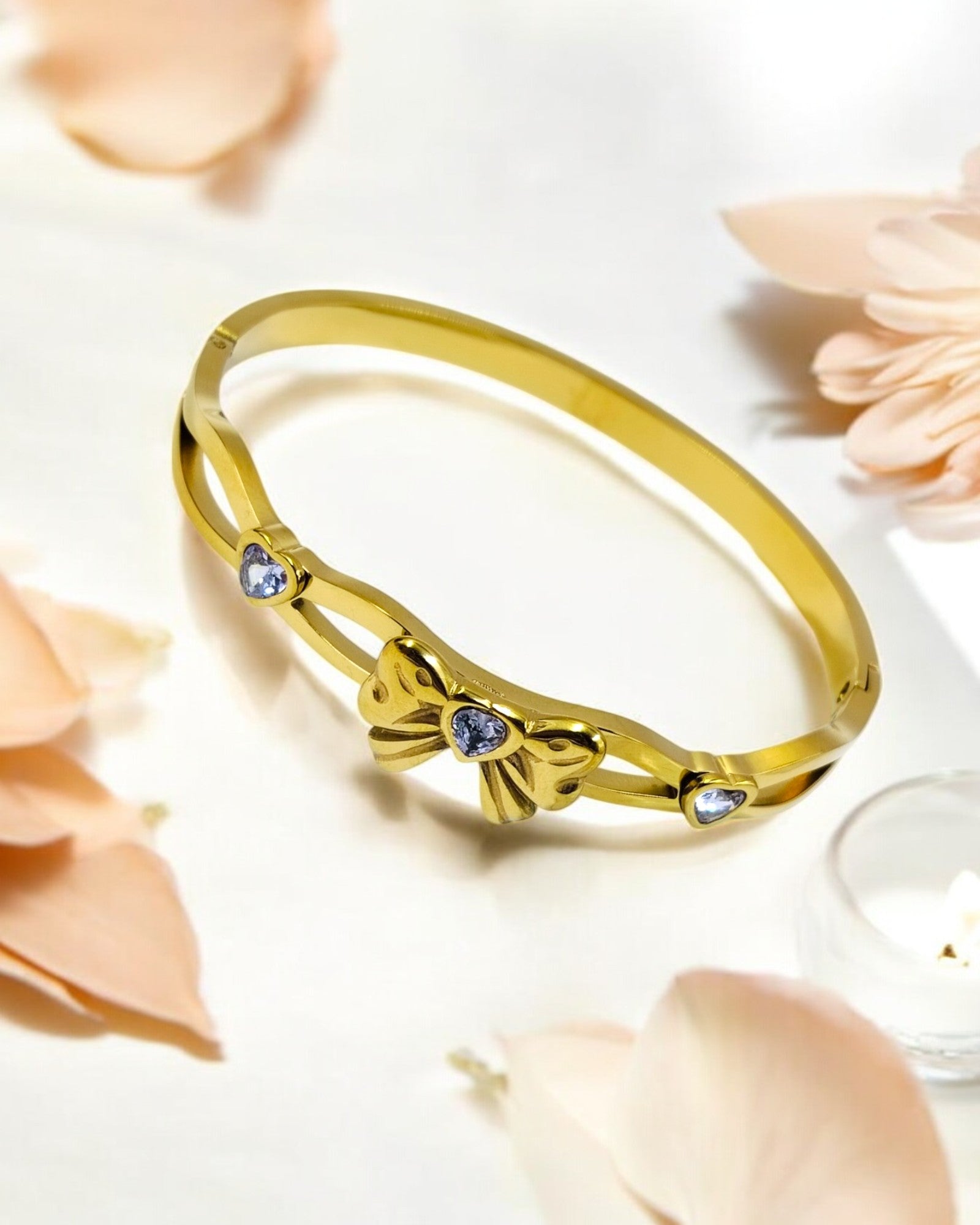 A gold-plated bracelet with a bowknot design and heart-shaped gemstone accents, displayed on a white surface with pink petals around it.