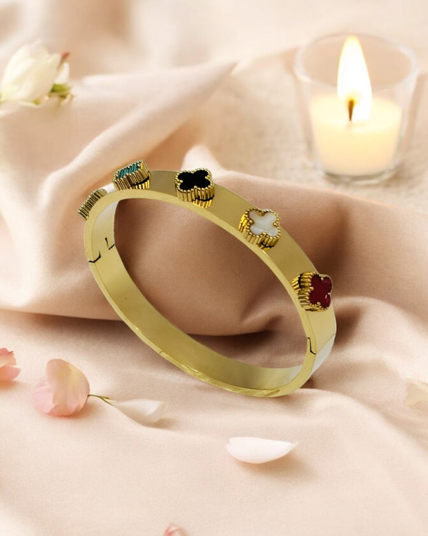 A gold-plated bangle bracelet with multi-colored clover accents, displayed on a satin fabric with a candle in the background.