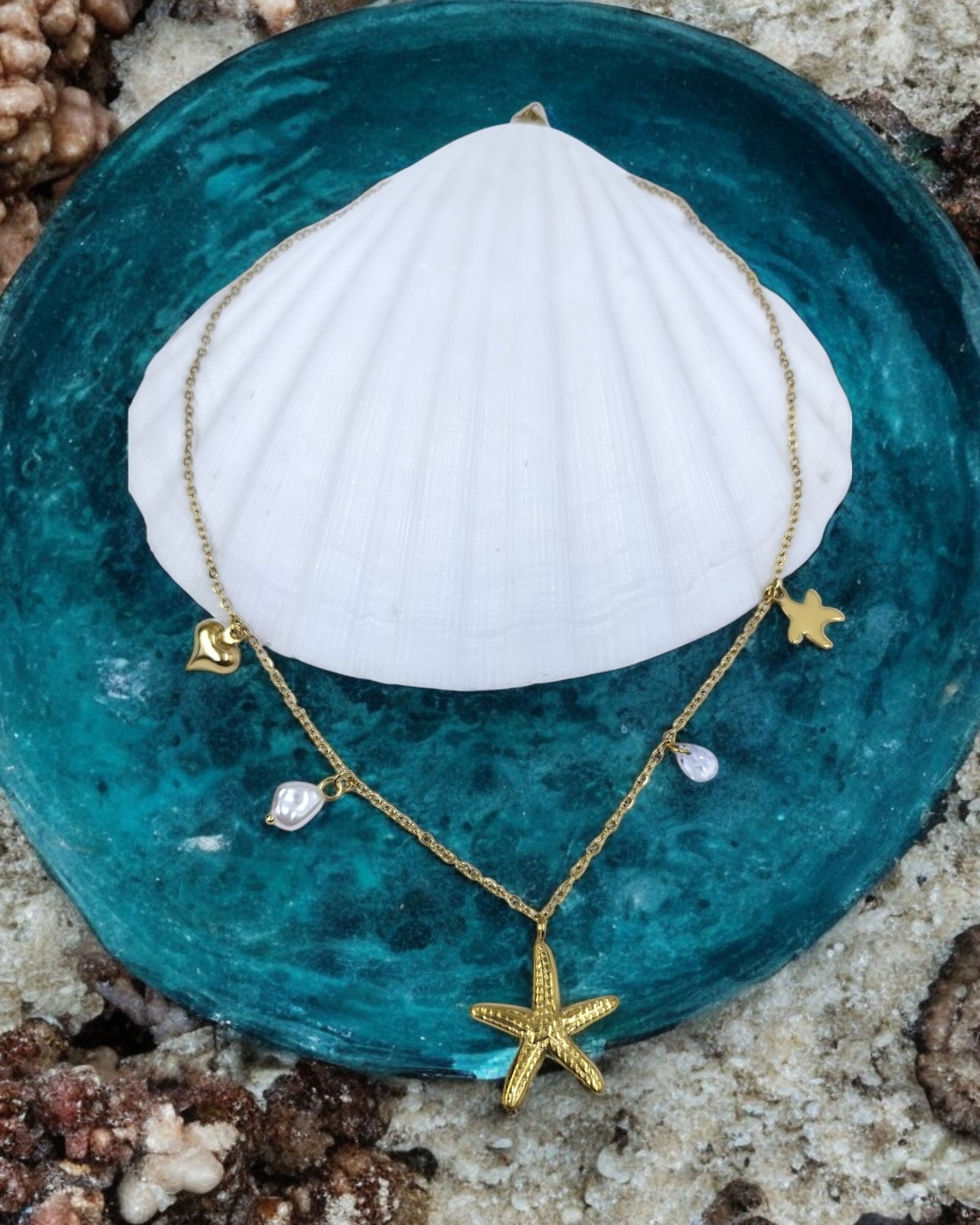 A gold starfish pendant necklace with small oceanic charms and pearl details, displayed on a blue dish with a white shell in the background.