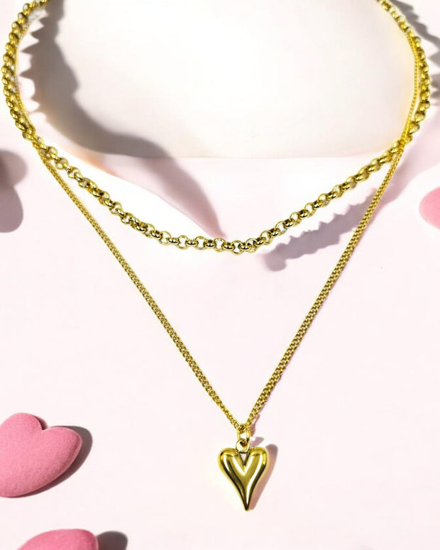 A gold-colored multilayer chain necklace with a heart pendant displayed against a white background with pink heart accents.