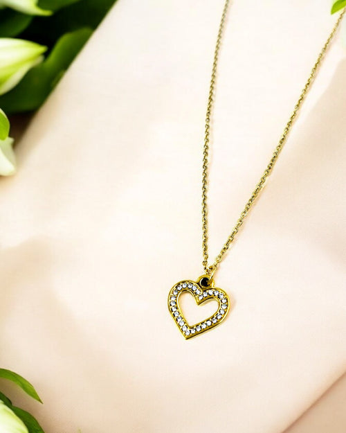 Gold chain necklace with an open heart-shaped pendant encrusted with sparkling white crystals, displayed on soft beige fabric with floral accents.