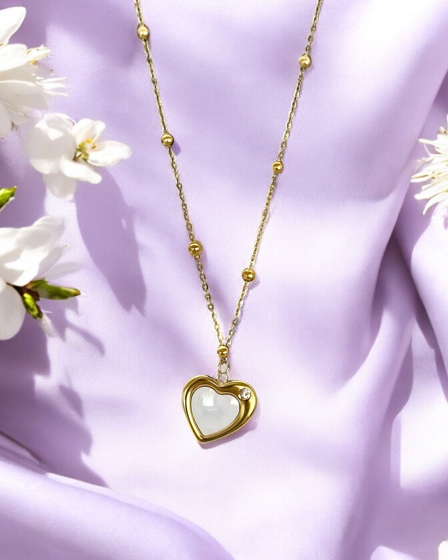 A gold-colored crescent heart pendant necklace with a white marble-like heart-shaped inset, displayed against a purple fabric background with white flowers.