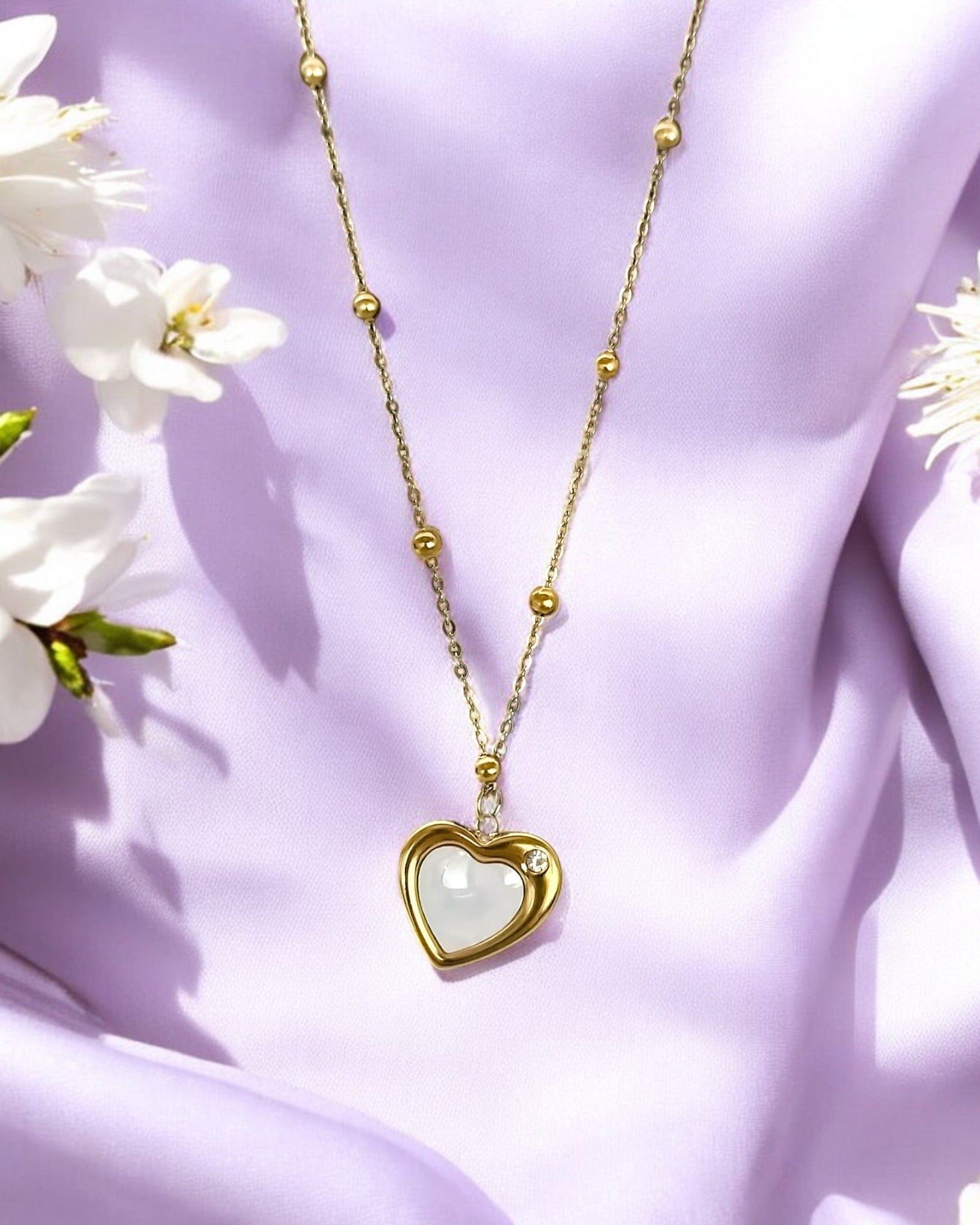 A gold-colored crescent heart pendant necklace with a white marble-like heart-shaped inset, displayed against a purple fabric background with white flowers.