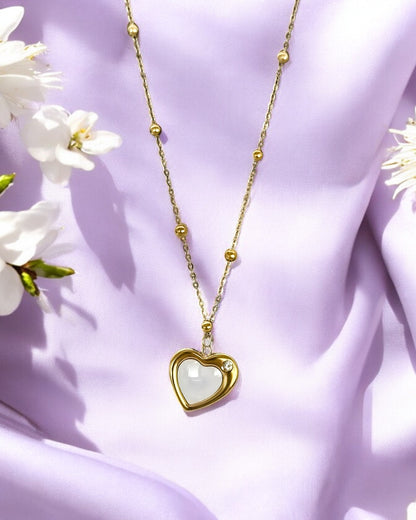 A gold-colored crescent heart pendant necklace with a white marble-like heart-shaped inset, displayed against a purple fabric background with white flowers.