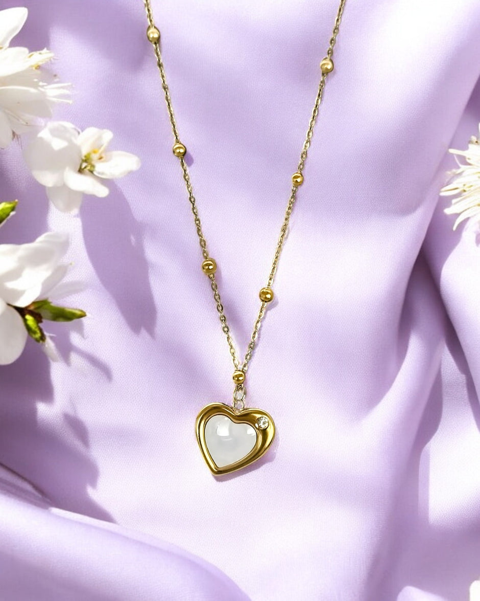 A gold-colored crescent heart pendant necklace with a white marble-like heart-shaped inset, displayed against a purple fabric background with white flowers.