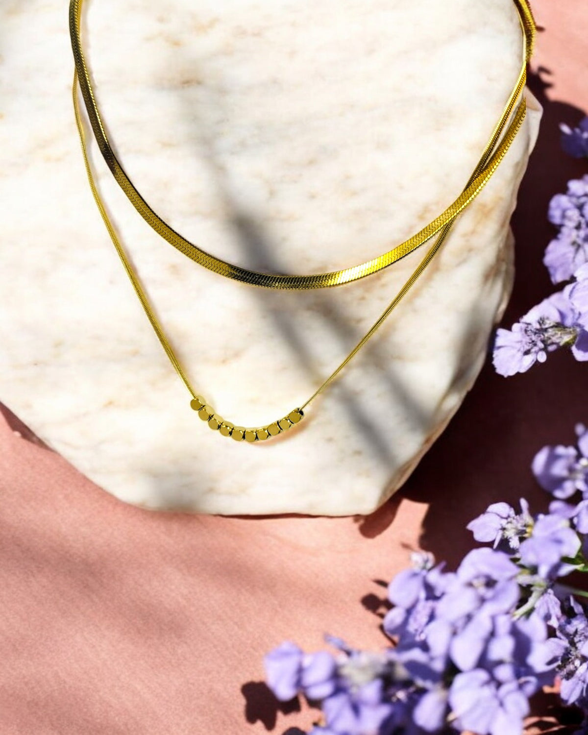 A gold-colored double layered square beaded chain necklace displayed on a white surface with purple flowers nearby.