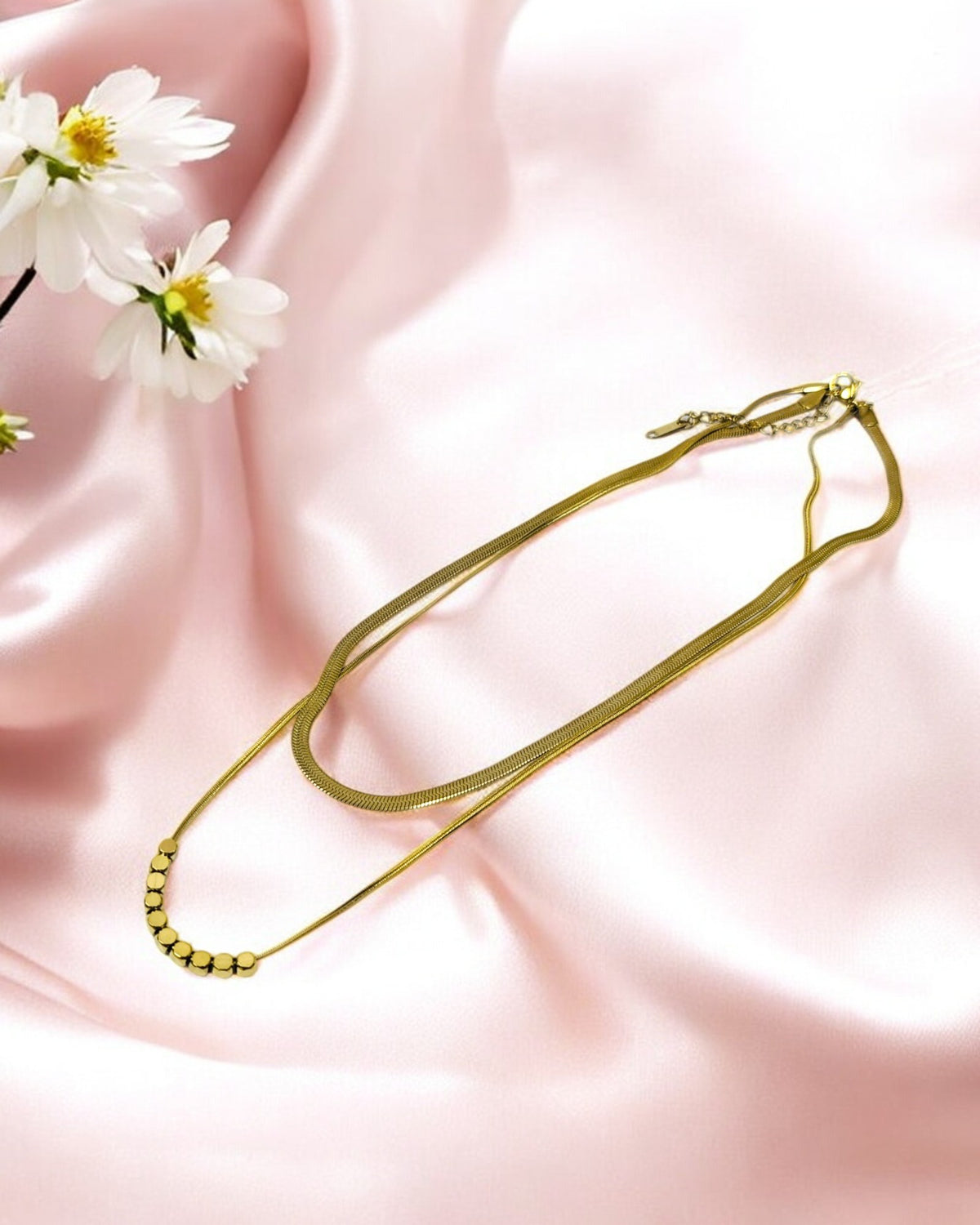 Elegant layered gold necklace with beaded detail and sleek flat chain, displayed on soft pink satin with white flowers.