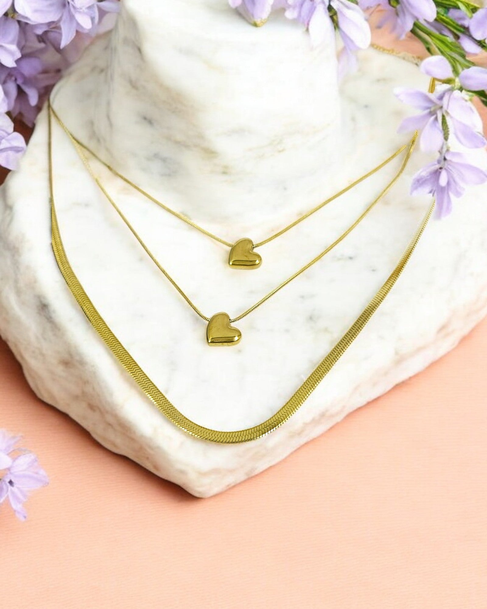A triple layered heart-themed chain necklace displayed on a marble surface with a soft pink background and small purple flowers around it.