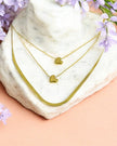 A triple layered heart-themed chain necklace displayed on a marble surface with a soft pink background and small purple flowers around it.