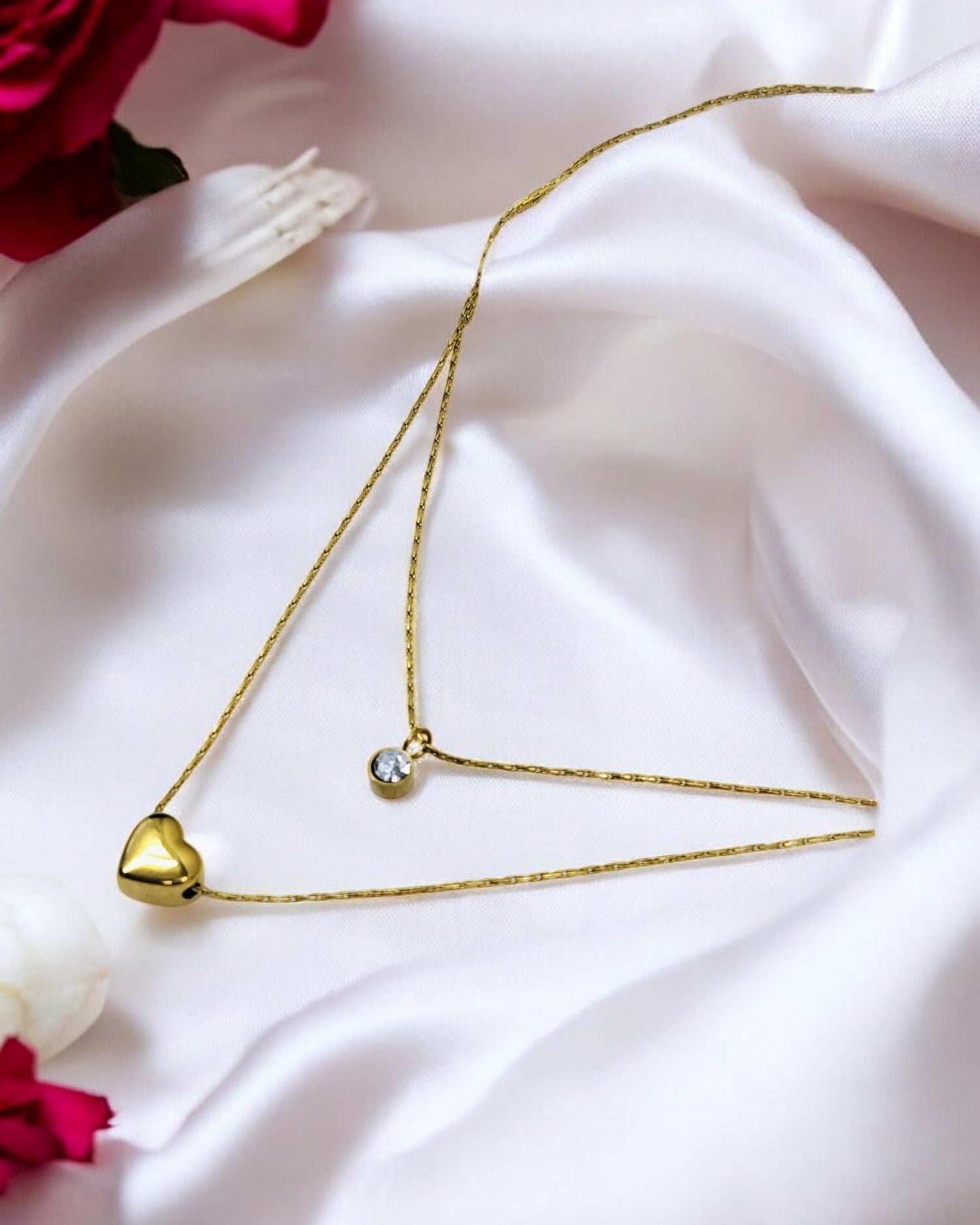 A gold-colored double layer chain necklace with a heart-shaped pendant displayed on a white fabric surface, possibly satin.