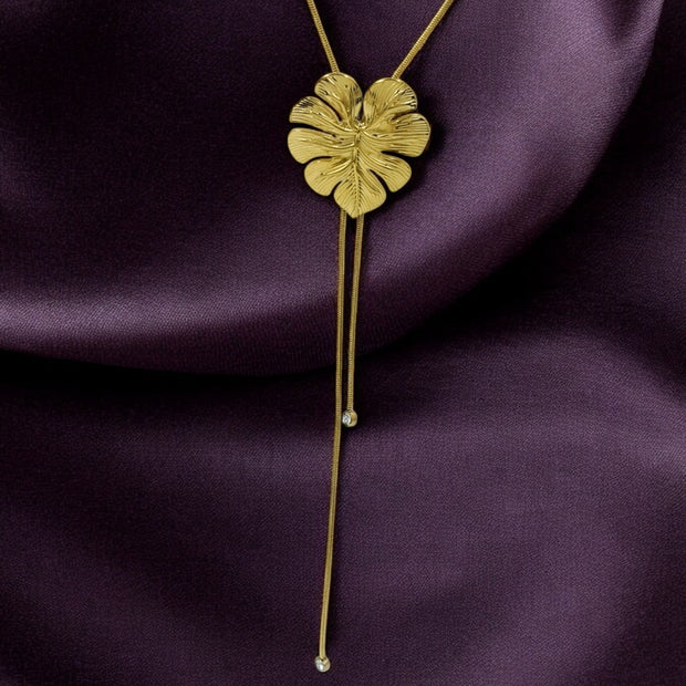 A gold-colored palm leaf pendant on a long Y chain necklace, displayed on a purple background.